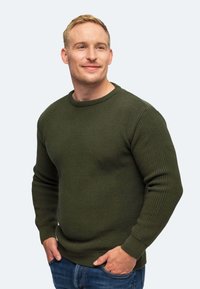 Grüner gestrickter Pullover mit Rundhalsausschnitt, strukturierter Stoff, gerippte Bündchen und Saum, kombiniert mit blauen Jeans, getragen von einem männlichen Model.