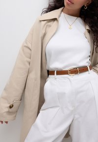 Beige trench coat with large collar, white t-shirt, brown leather belt with gold buckle, and loose white trousers. Subtle texture and tailored fit.