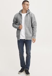 Grå hettegenser med glidelås og kengurulomme, snor i hetta, kombinert med en hvit T-skjorte og blå jeans, samt svarte sneakers.