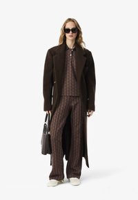 Brauner Wollmantel, gemustertes braunes Outfit mit geometrischem Design und weißen Schuhen. Model hält eine dunkle Handtasche und trägt schwarze Sonnenbrille.