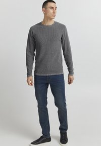 Maglietta grigia a maniche lunghe con un motivo testurizzato, colletto a giro, abbinata a jeans blu e sneakers nere, mostrata su un modello in piedi.
