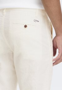 Beige linbukser med en glat tekstur, med en baglomme med en brun knap og et synligt logo-label ovenfor.