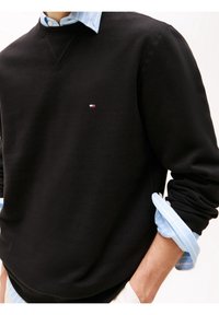 Felpa nera con scollo rotondo, caratterizzata da un piccolo logo. Indossata sopra una camicia a righe blu chiaro con le maniche arrotolate.
