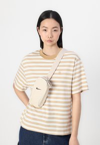 Beige crossbody bag with Nike logo, featuring a textured finish and white accents, worn over a striped beige and cream t-shirt.