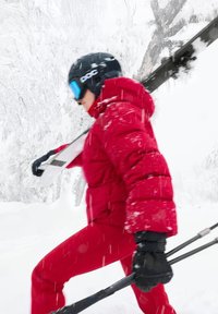 Röd puffig skidjacka och matchande byxor, svarta handskar och hjälm med blåa goggles; bär skidstavar i en snötäckt miljö.