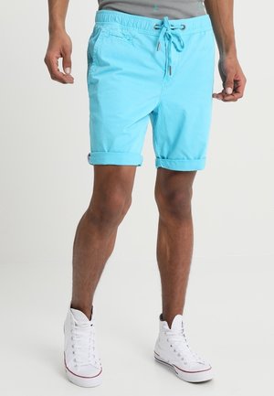Turkusowe szorty z elastycznym paskiem i sznurkiem, z podwiniętymi mankietami i bocznymi kieszeniami. Noszone z białymi trampkami wysokimi.