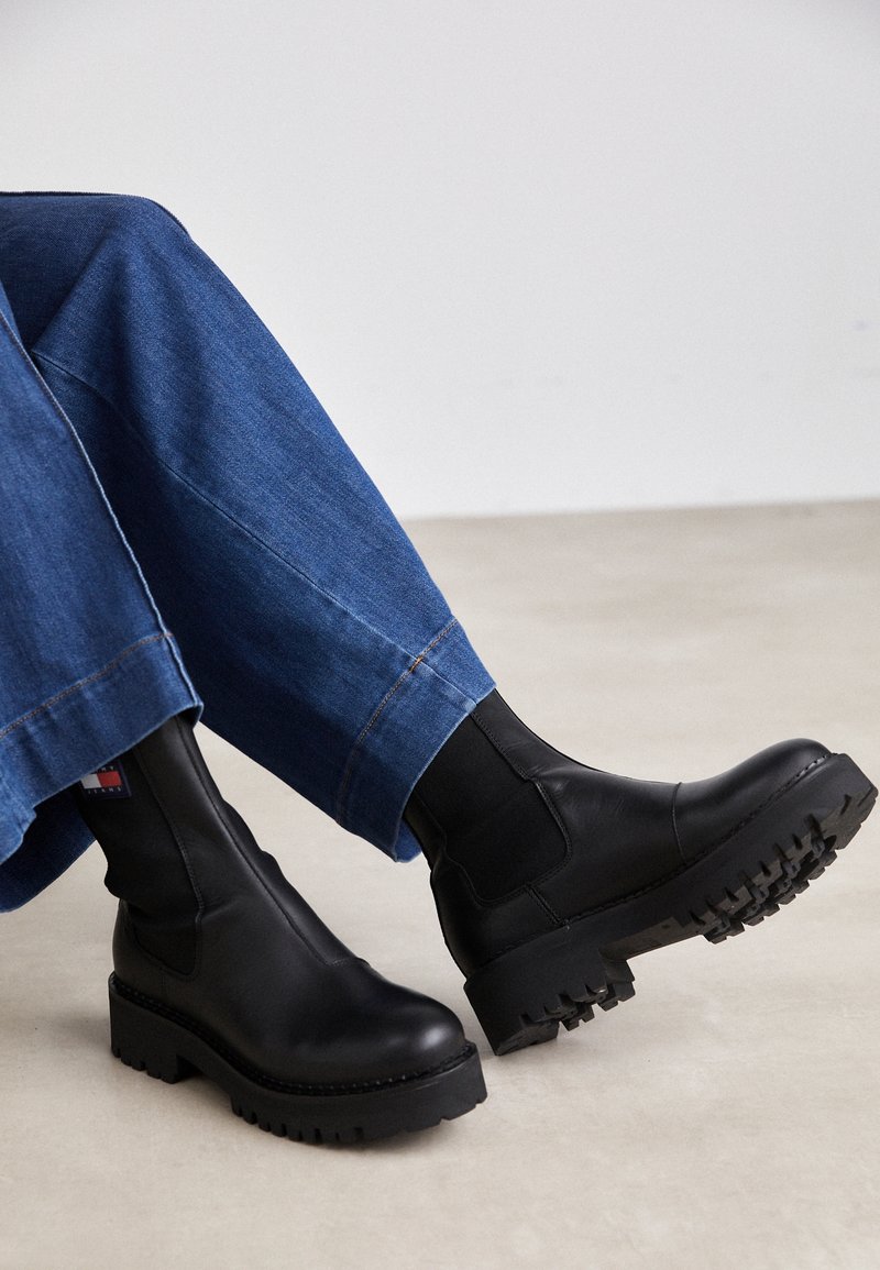 Vue en longueur des jambes d'une personne portant un pantalon en denim bleu et des bottines noires à semelles épaisses avec des panneaux élastiques sur les côtés.
