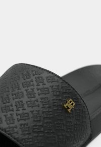 Sandale noire à glissière avec un dessus texturé à motif logo répété et un accent doré. Semelle lisse avec une légère courbe pour plus de confort.