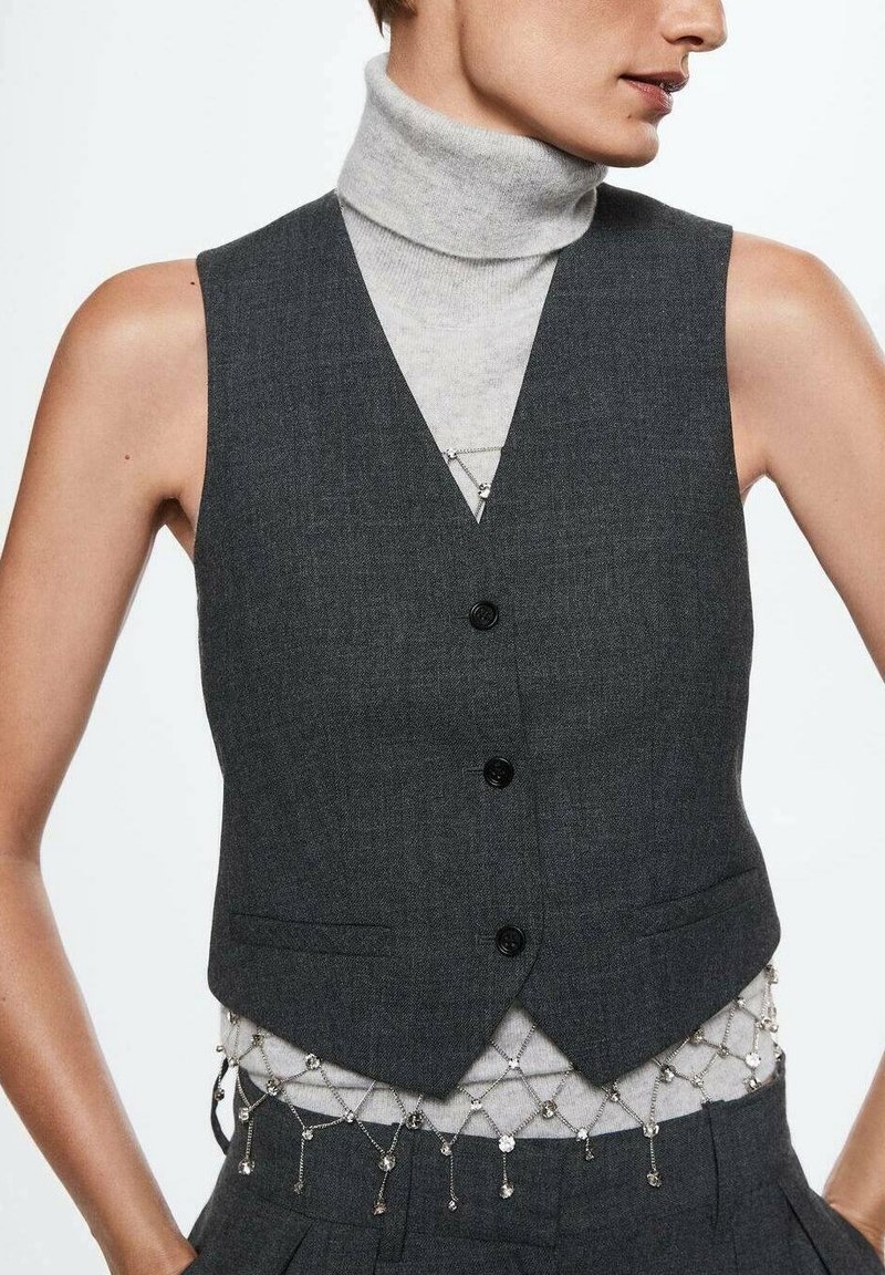Femme portant un gilet boutonné gris foncé sur un col roulé gris clair et une ceinture chaîne argentée avec des breloques pendantes autour de la taille.