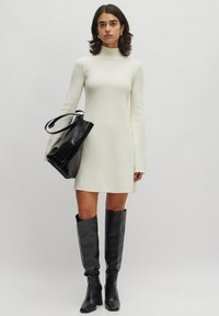 Weißes langärmliges Kleid mit hohem Ausschnitt, kombiniert mit hohen schwarzen Lederstiefeln und einer glänzenden schwarzen Handtasche. Schlichtes, minimalistisches Design.