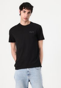 Schwarzes Baumwoll-T-Shirt mit kurzen Ärmeln, ausgestattet mit einem kleinen weißen "TOMMY JEANS"-Logo auf der Brust. Getragen mit hellblauen Jeans.