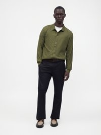 Homme portant une chemise kaki boutonnée, un sous-vêtement blanc, un pantalon noir, une ceinture marron et des chaussures bicolores marron et beige, debout sur fond blanc.