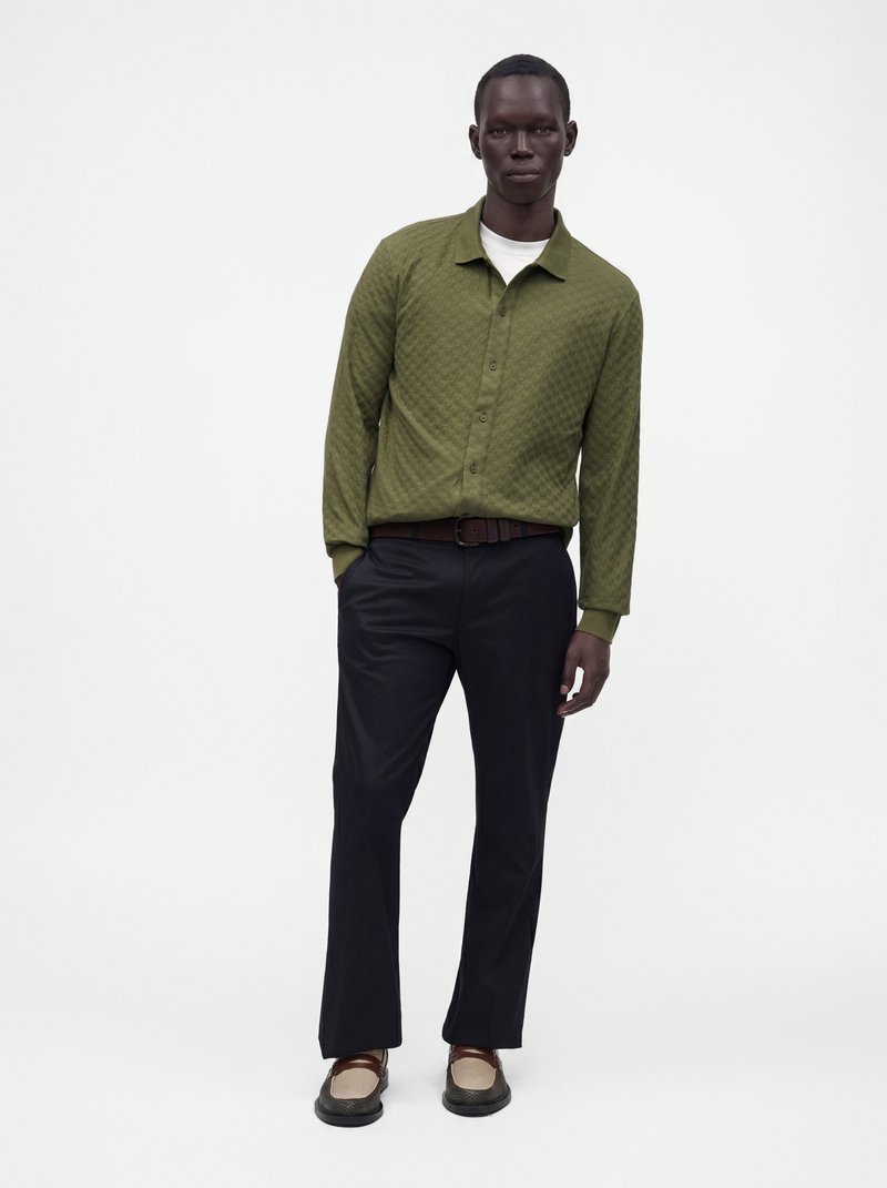 Homme portant une chemise kaki boutonnée, un sous-vêtement blanc, un pantalon noir, une ceinture marron et des chaussures bicolores marron et beige, debout sur fond blanc.