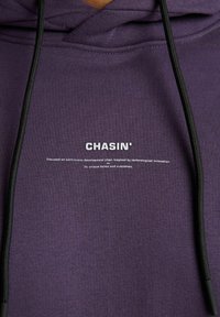 Sudadera púrpura con cordón, un logo blanco "CHASIN'" en el frente y texto relacionado con el desarrollo continuo y la innovación.