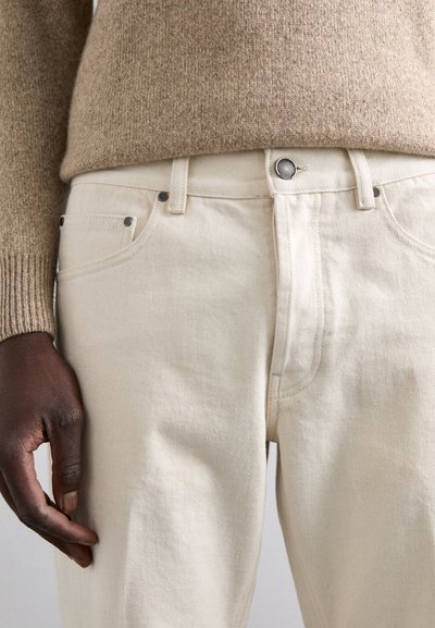 Jeans de algodón beige con corte clásico, que cuentan con un cierre de botón plateado, bolsillos laterales y sutiles detalles de costura. Textura suave visible.