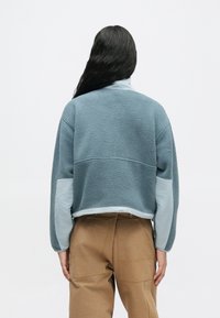 Άτομο με μακριά μαύρα μαλλιά, φορώντας μια κοντή μπλε φλις ζακέτα και καφέ παντελόνι ψηλόμεσο, στέκεται με την πλάτη στραμμένη στη λευκή φόντο.