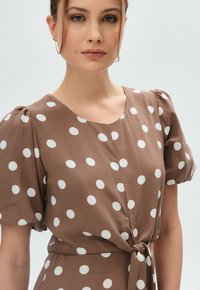 Braunes Kleid mit Polka-Dots, kurzen Puffärmeln, einem runden Ausschnitt und einer taillierten Bindung. Glatte Stoffstruktur; großes Muster mit weißen Punkten.