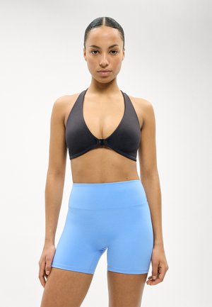 Ženska stoji obrnjena naprej, oblečena v črni športni nedrček s sprednjo zaponko in svetlo modre visoko pasne športne kratke hlače.