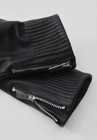 Ärmel einer schwarzen Lederjacke mit gerippten Details und silbernen Metallreißverschlüssen vor neutralem Hintergrund.