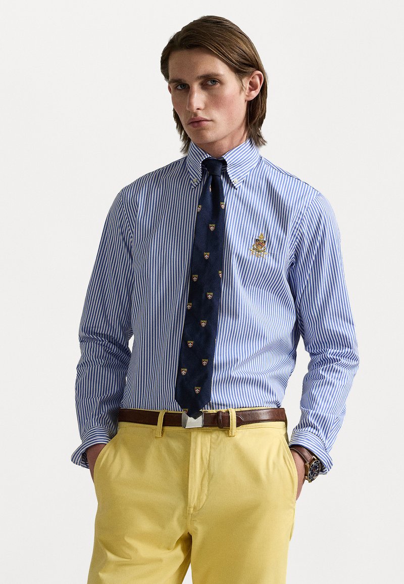 Prugasta plava i bijela košulja s dugim rukavima, uz tamnoplavu kravatu s malim grbovima, uparena s žutim hlačama i smešnim remenom.