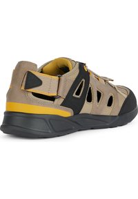 Zapato exterior beige y negro con acentos amarillos, que cuenta con paneles de malla, un cuello acolchado y una suela de goma texturizada. Cierre de correa ajustable.