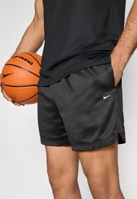Μαύρο αθλητικό σορτς Nike με υφή από δίχτυ, με πλευρικές σχισμές, μικρό λευκό λογότυπο στο στρίφωμα και ένα μπάσκετ στο άλλο χέρι.