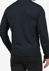 Homme portant un pull bleu marine ajusté et un jean noir, vu de dos avec une main détendue le long du corps.