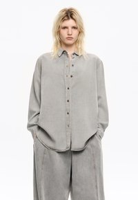 Helle graue Denim-Bluse mit Knopfleiste, Kragen und langen Ärmeln, mit einem lässigen Schnitt und sichtbaren Nahtdetails.