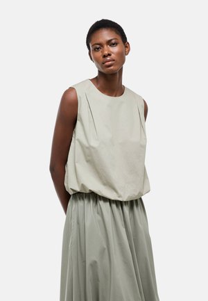 Femme portant un haut sans manches ample de couleur gris clair avec des plis et une jupe taille haute assortie, debout devant un fond blanc uni.