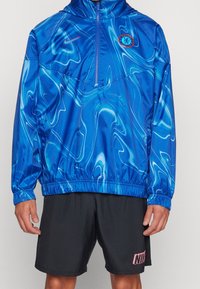 Mężczyzna ubrany w niebieską kurtkę Chelsea Football Club z falistym wzorem i czarne szorty Nike z różowym logo, stojący na jednolitym tle.
