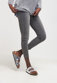 Hopeiset sandaalit, joissa on kaksi säädettävää remmiä, yhdistettynä harmaisiin skinny-farkkuihin ja teksturoituun harmaaseen neuleeseen, valkoista taustaa vasten.