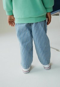 Blau-weiß gestreifte Hosen mit lockerem Schnitt, kombiniert mit einem grünen Sweatshirt. Sichtbare weiße Turnschuhe mit roten Akzenten.