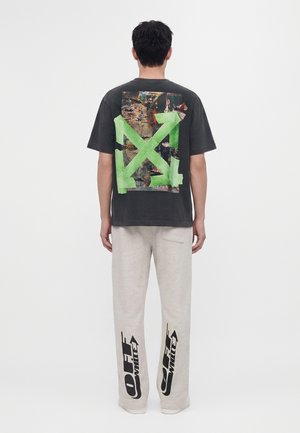 Persoon die een zwart t-shirt draagt met een groot groen pijltje ontwerp en een bedrukte collage op de rug, gecombineerd met lichte broek met zwarte Off-White-logo's.