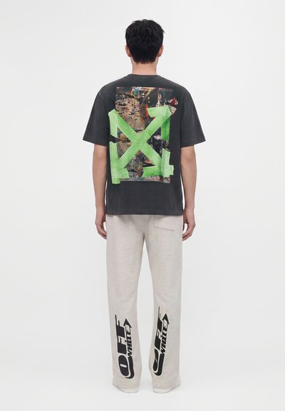 Person wearing a black t-shirt with a large green arrow design and printed collage on the back, paired with light pants featuring black Off-White logos.