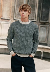 Knitted sweater featuring a herringbone pattern in navy and cream colors, with a cream ribbed neckline and cuffs, paired with dark jeans.