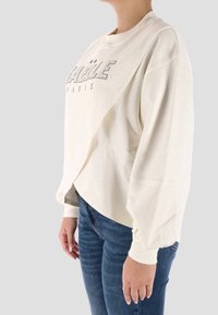 Felpa color crema con polsini a costine e vestibilità ampia, con la scritta "PARIGI" in strass sul davanti. Abbinata a jeans blu.