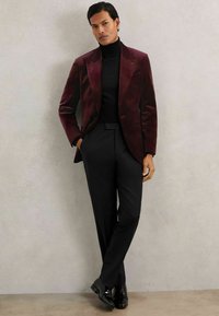 Blazer di velluto bordeaux sopra un dolcevita nero, abbinato a pantaloni neri e scarpe nere lucide, su uno sfondo neutro.