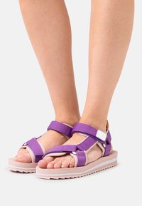 Violet kangas sandaalit säädettävillä hihnoilla, valkoisilla yksityiskohdilla ja teksturoidulla vaaleanpunaisella pohjalla. Avoin kärki tarjoaa mukavuutta vaimentavalla sisäpohjalla.
