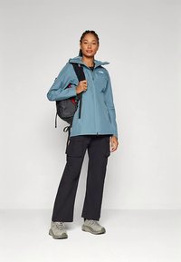 Niebieska wodoodporna kurtka z kapturem, zamkiem z przodu i kieszenią na piersi. W zestawie z czarnymi spodniami typu cargo i szarymi butami trekkingowymi. Plecak zarzucony na jedno ramię.