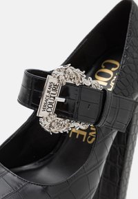 Escarpin en cuir verni noir avec texture crocodile, matériel de boucle en argent et détails de marque. Design à bout fermé et avec sangle à la cheville.