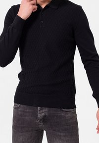 Pull polo en maille noire avec un motif texturé en losanges, à manches longues et col boutonné, associé à un jean gris foncé.