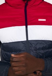 Polstret jakke med et farveblokdesign i rød, hvid og marineblå; har hætte, lynlås lukning og logo på brystet. Struktureret stof.