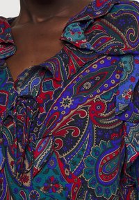 Mångfärgad skjorta med paisleymönster i röda, blå och gröna nyanser; volangkrage och slipsdetalj vid halsringningen.