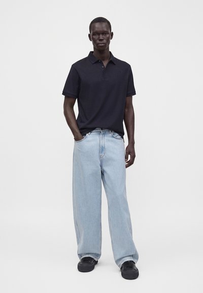 Homme portant un polo noir à manches courtes, un jean bleu clair à jambes larges avec les mains dans les poches et des baskets noires, debout devant un fond uni.