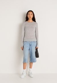 Svetlošedý svetrový top s dlhým rukávom a okrúhlym výstrihom, skombinovaný s svetlo denimovými culottes a bielymi teniskami. Drží čiernu tašku s bielymi bodkami.