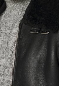 Veste noire en cuir avec un col en shearling noir texturé, une fermeture éclair argentée et un pull gris contrastant en tricot en dessous.