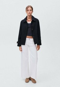 Jeune femme portant une veste noire courte, un crop top noir, un pantalon large blanc, des chaussures marron et un collier de perles rouges, se tenant devant un fond gris.