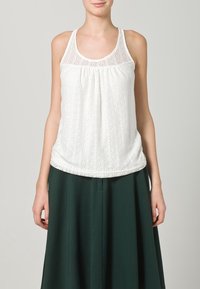 Top in pizzo bianco con scollo rotondo, vestibilità svasata e intricate fantasie circolari. Abbinato a una gonna verde scuro.