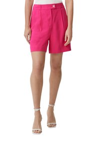 Donna che indossa pantaloni a vita alta di un brillante colore rosa, con chiusura a bottoni e zip frontale, abbinati a tacchi white con cinturino alla caviglia e una maglietta bianca.