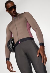 Cykeltrøje i brun og pink med lynlås foran. Tæt siddende design, lange ærmer og kontrasterende detaljer; parret med mørke cykelshorts og en hjelm.